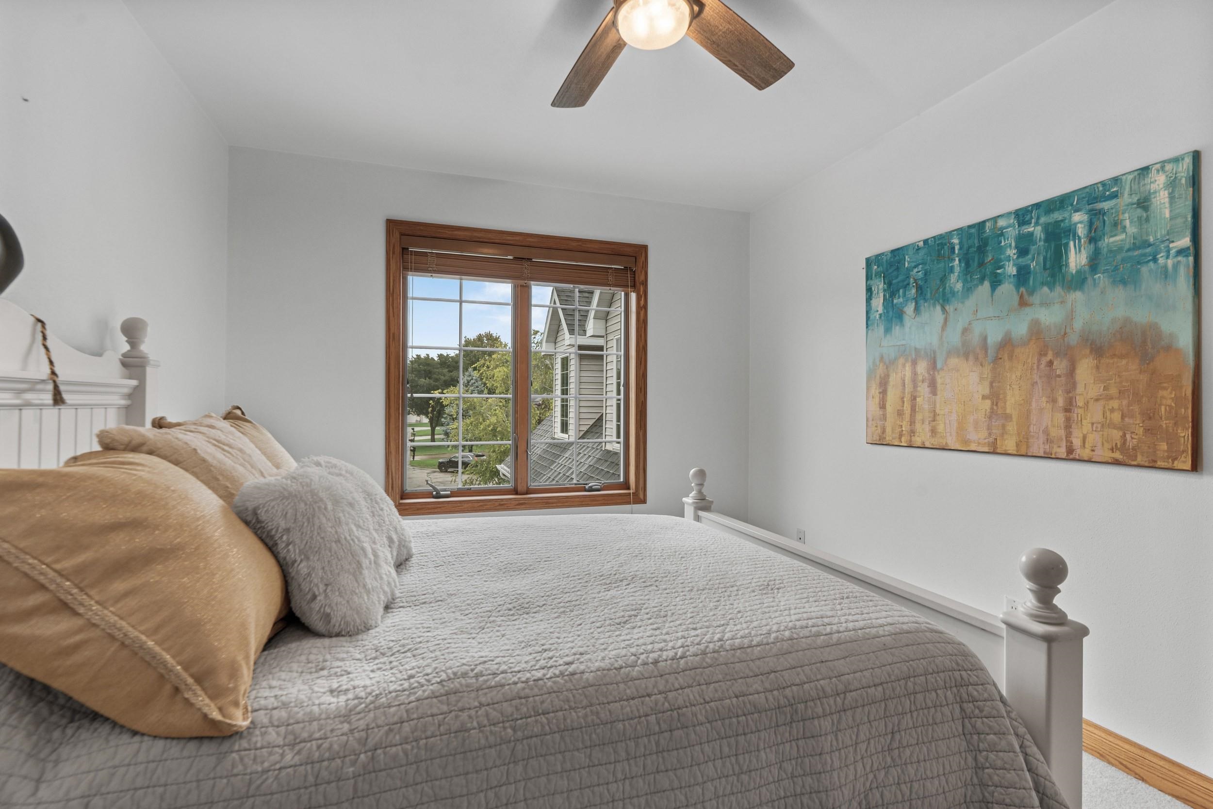 Bedroom with a gray quilted bed, tan pillows, and a wood-trimmed window overlooking trees.