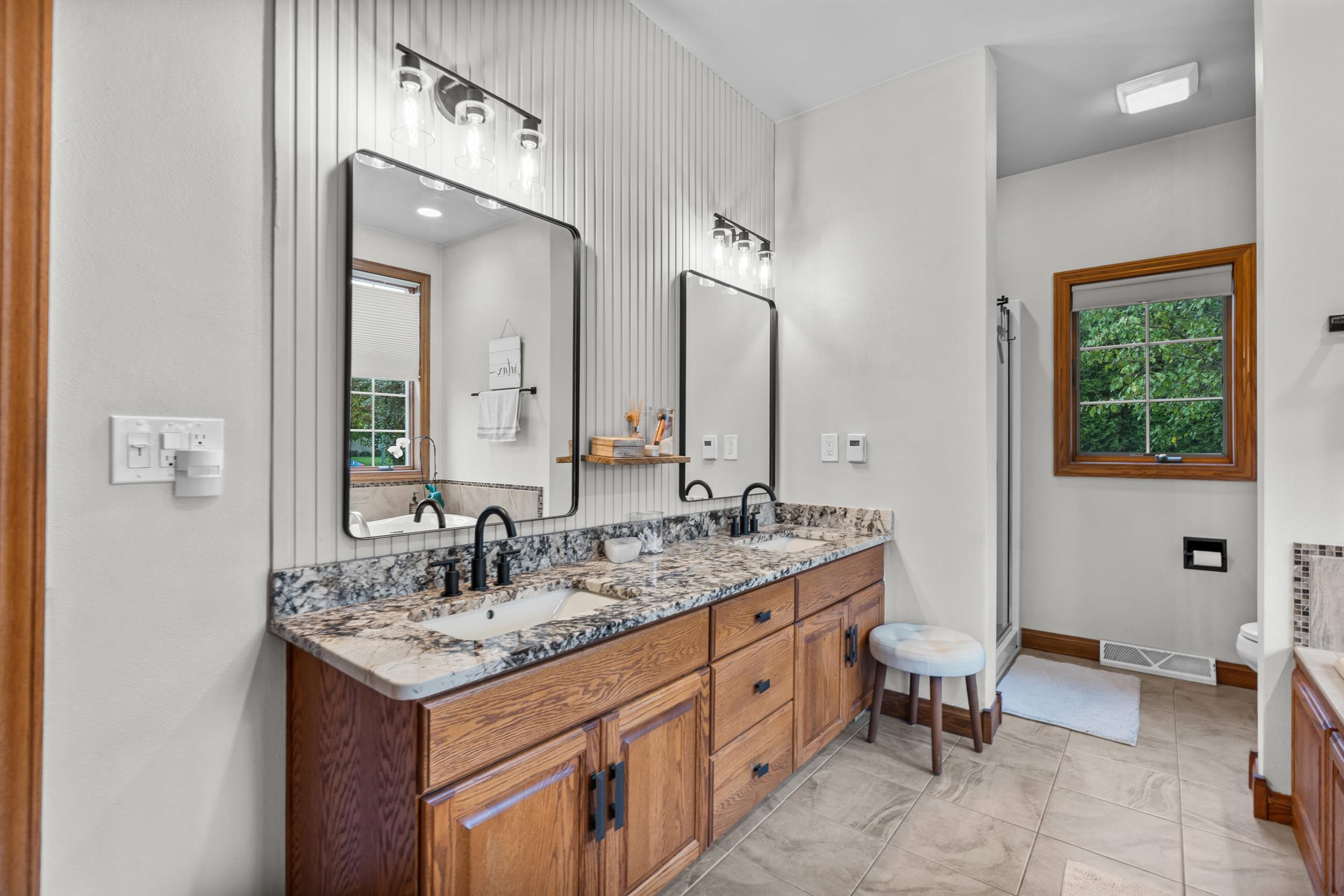 Bathroom with a double-sink wood vanity, stone counter, paneled wall, and wood-trimmed window.