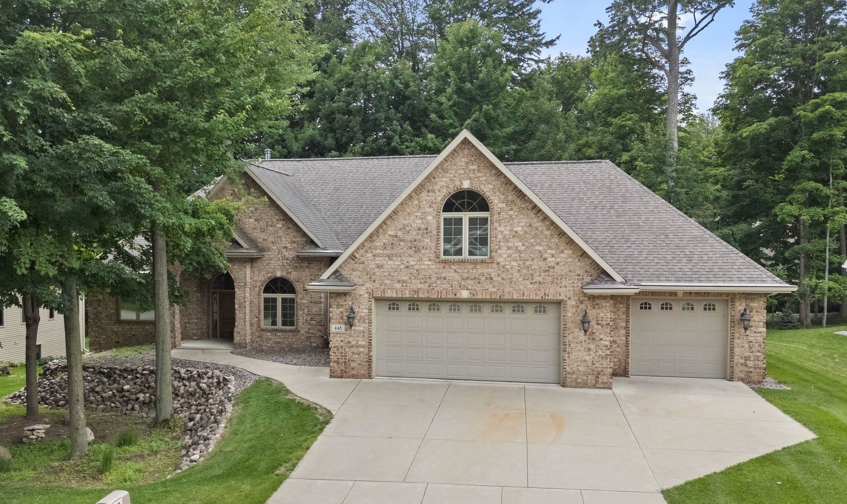 Brick house with a steep gray roof, arched upper window, and three-car garage beneath mature trees.