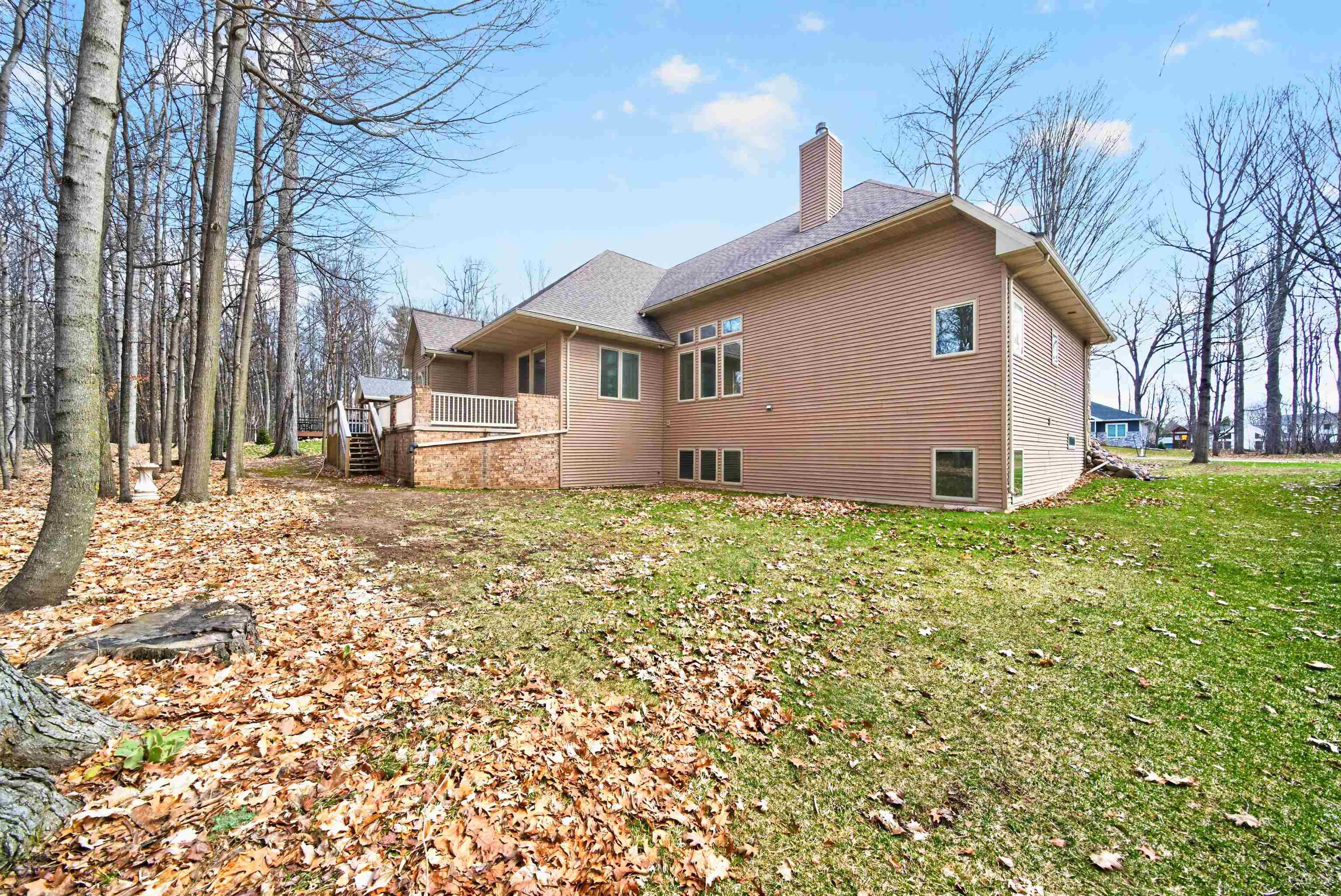 Rear of a brown-sided house with a raised deck and brick foundation beside a leaf-covered wooded yard.