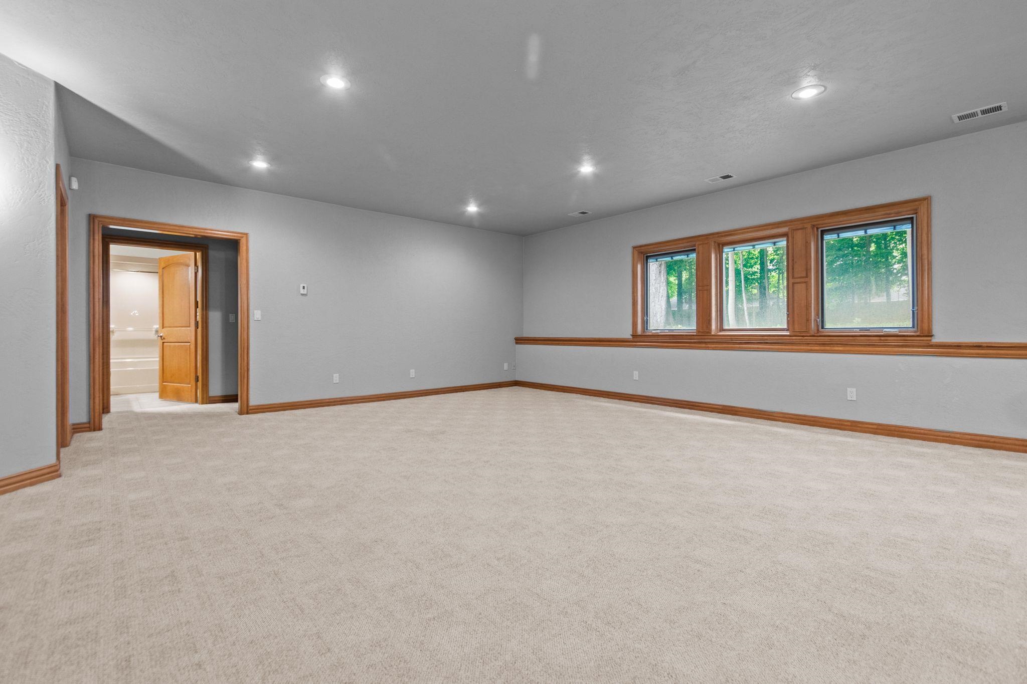 Empty carpeted room with wood-trimmed windows, recessed lights, and an open doorway.
