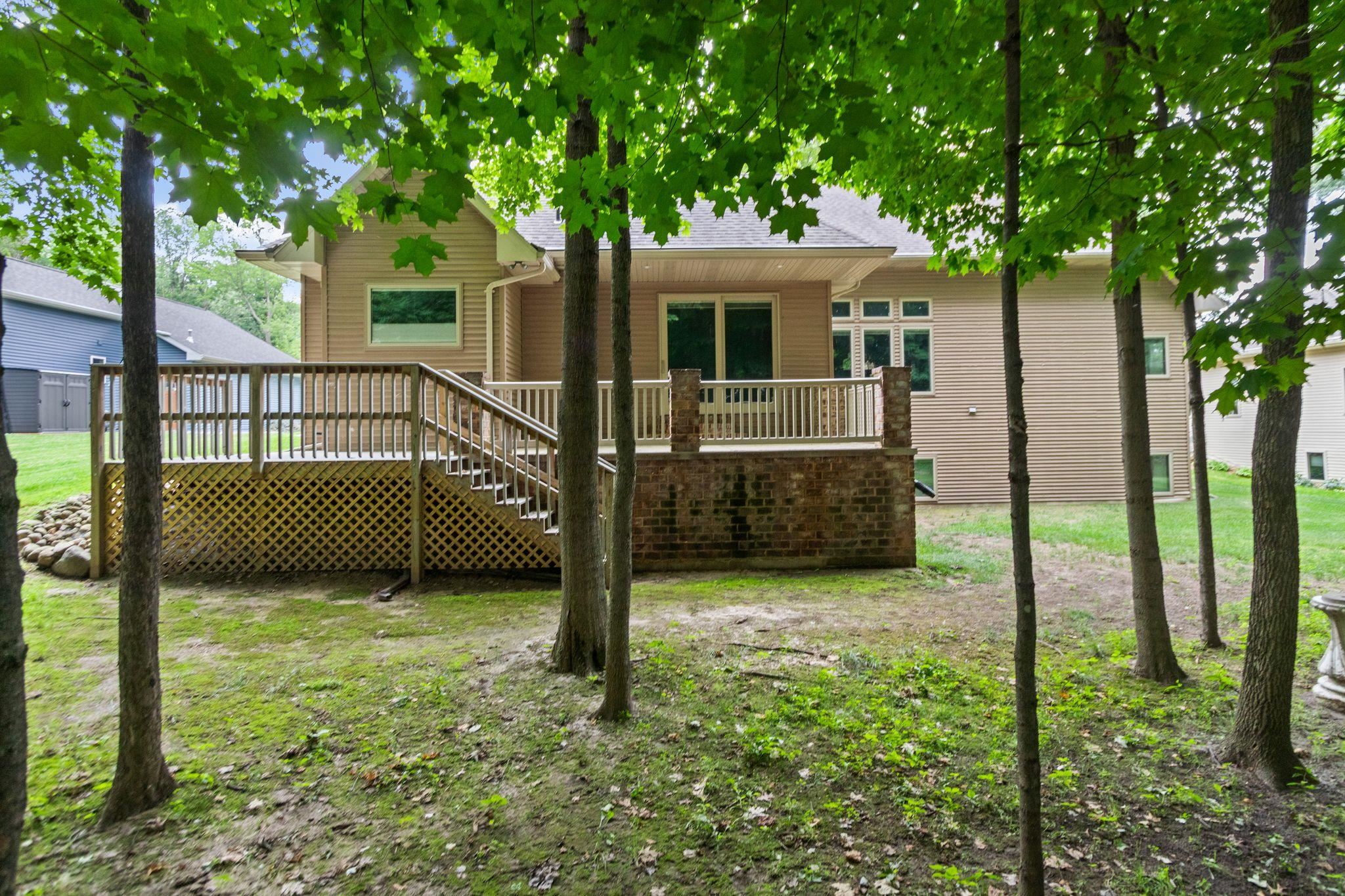 Rear of a tan-sided home with a raised deck and brick base, shaded by mature trees.