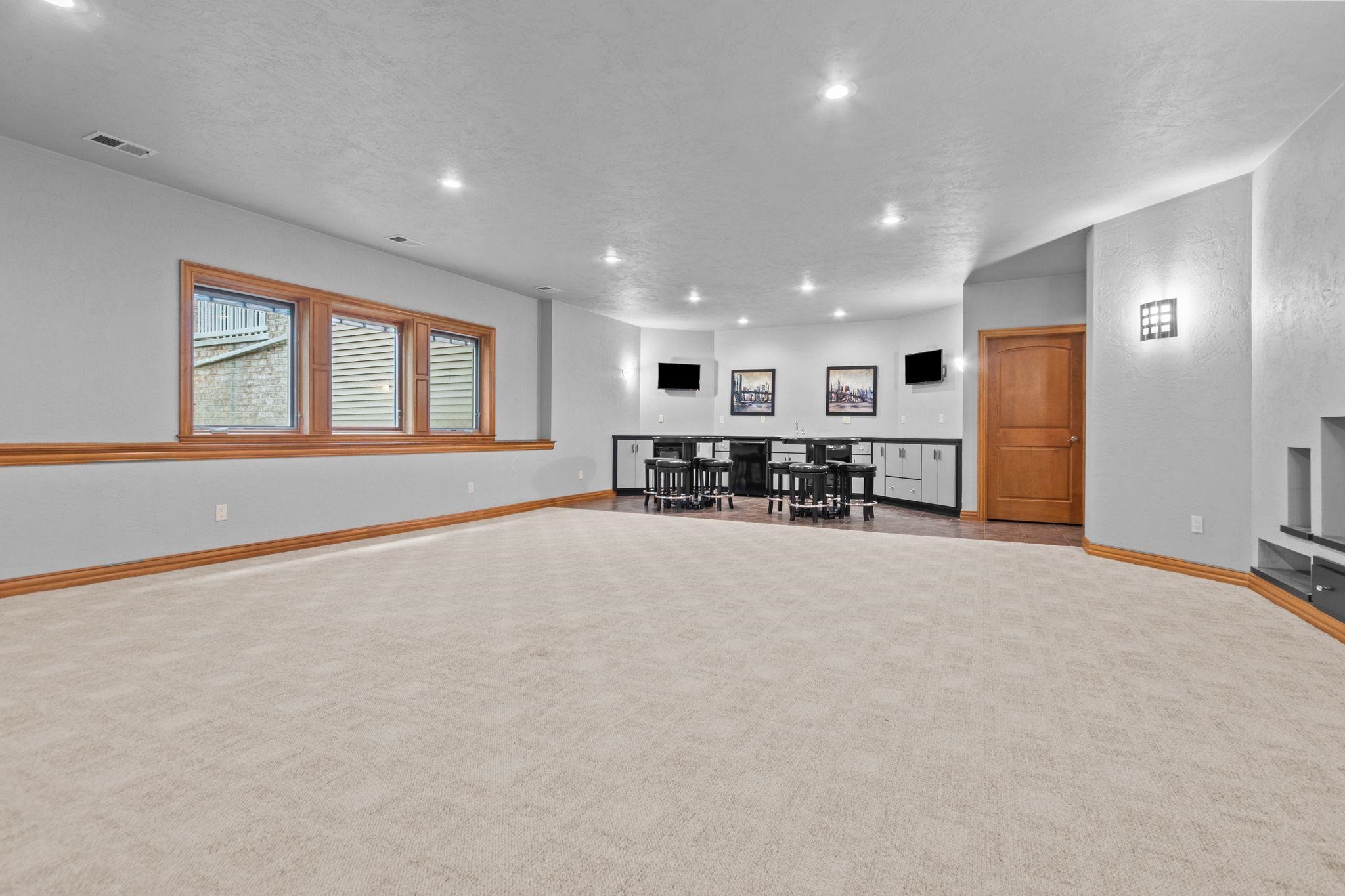 Carpeted basement recreation room with a built-in bar, black stools, and wood-trimmed windows.