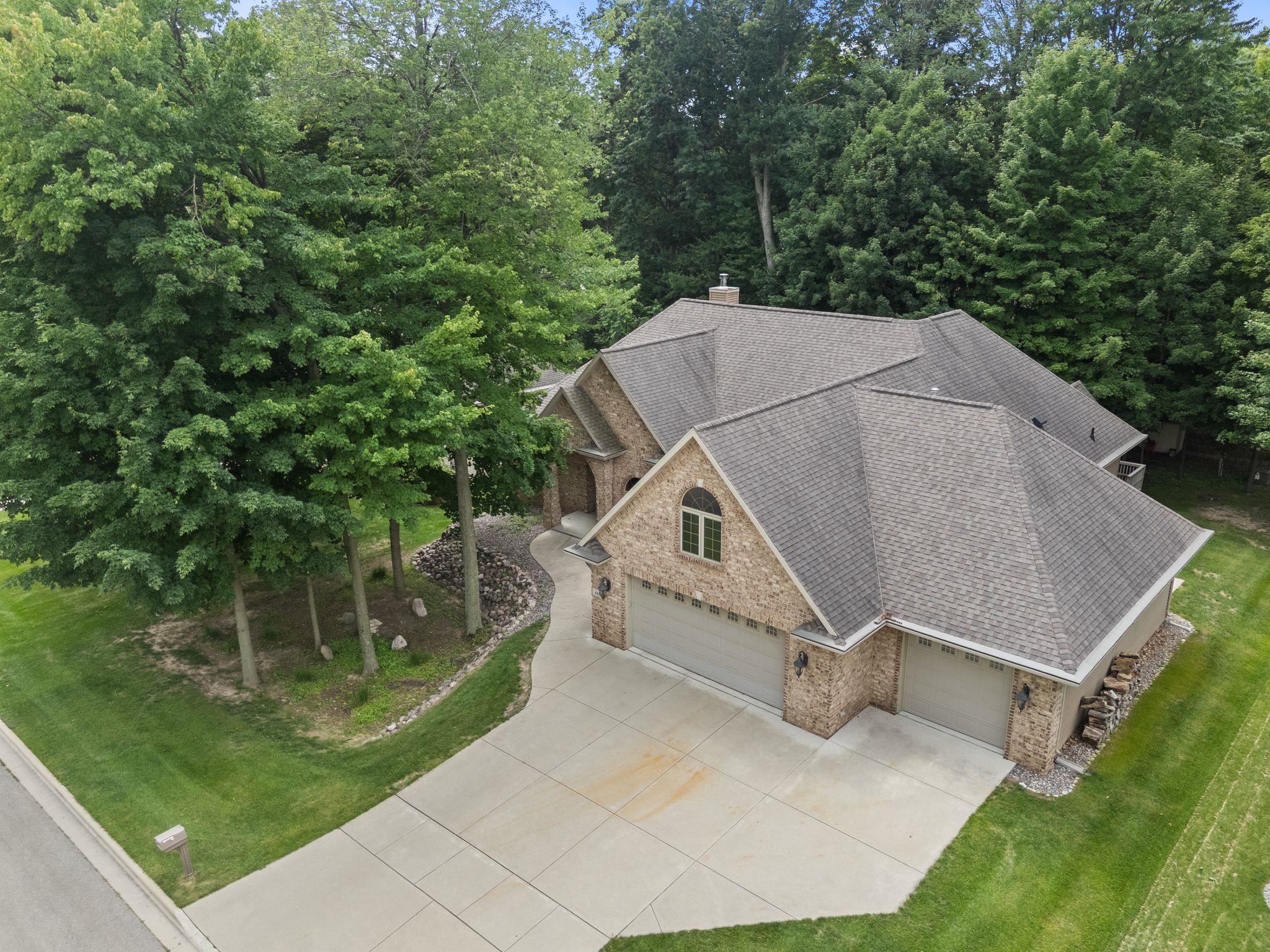 Aerial view of a brick home with multiple garage doors, a wide driveway, and mature trees.