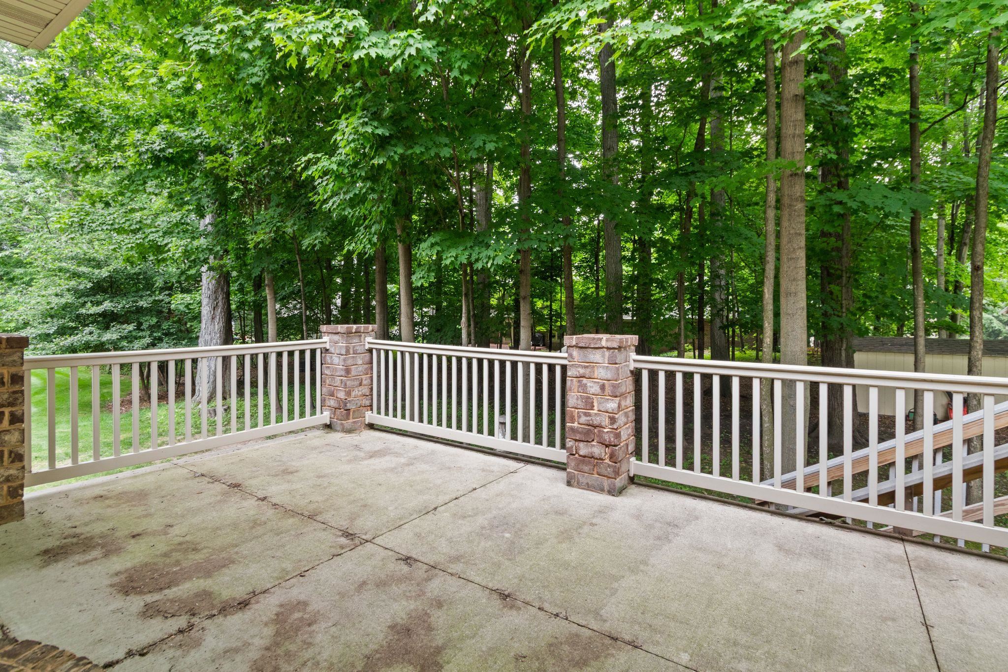Concrete deck with white railing and brick posts overlooking a leafy wooded backyard.