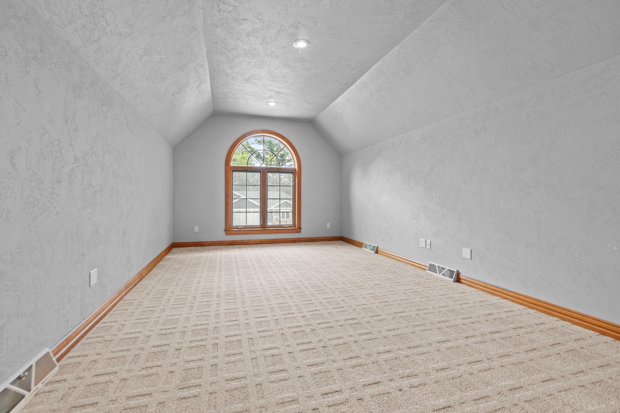 Empty carpeted room with a vaulted ceiling and a large arched, wood-trimmed window.