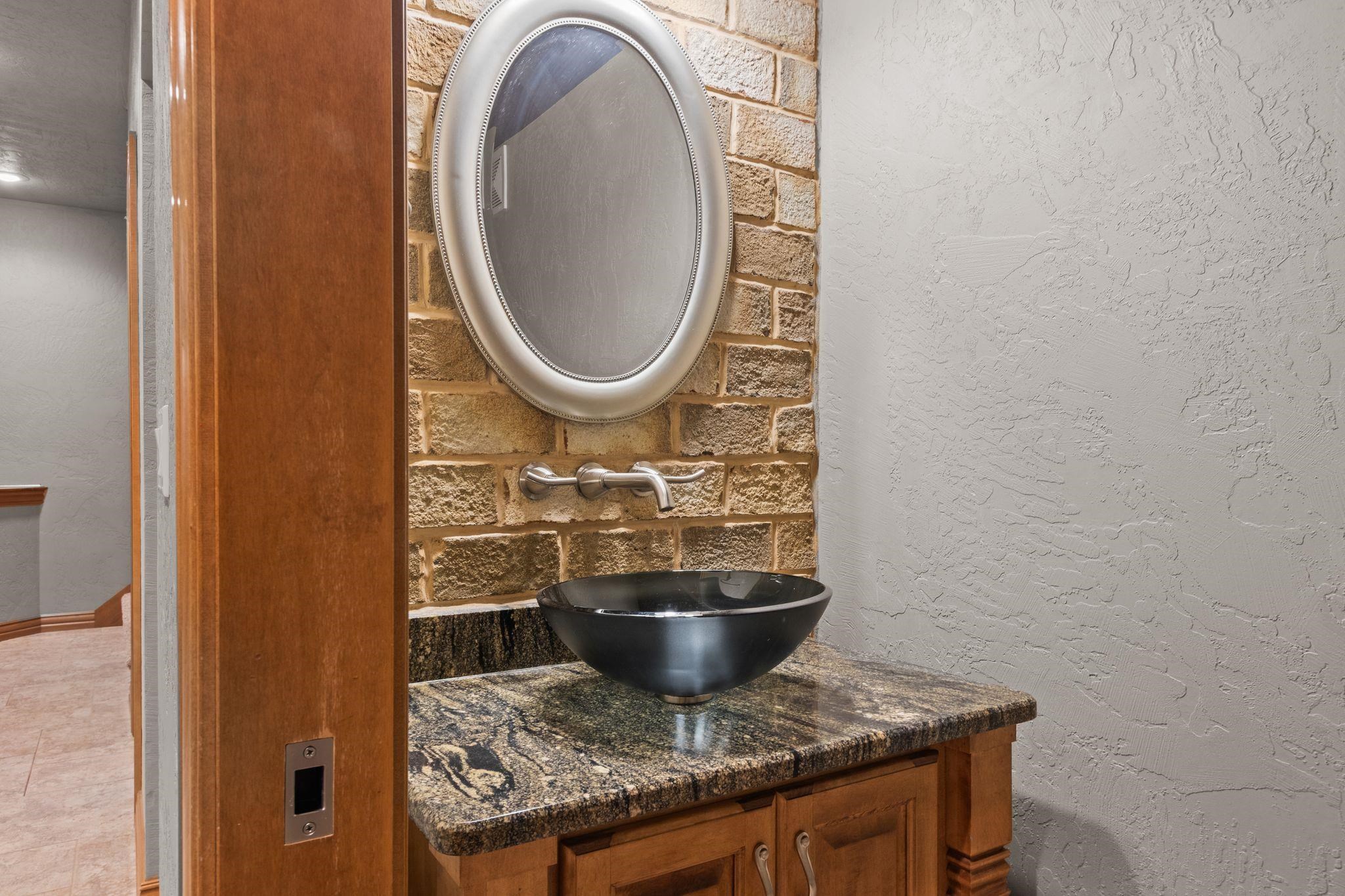 Bathroom vanity with a black vessel sink, granite countertop, brick accent wall, and oval mirror.