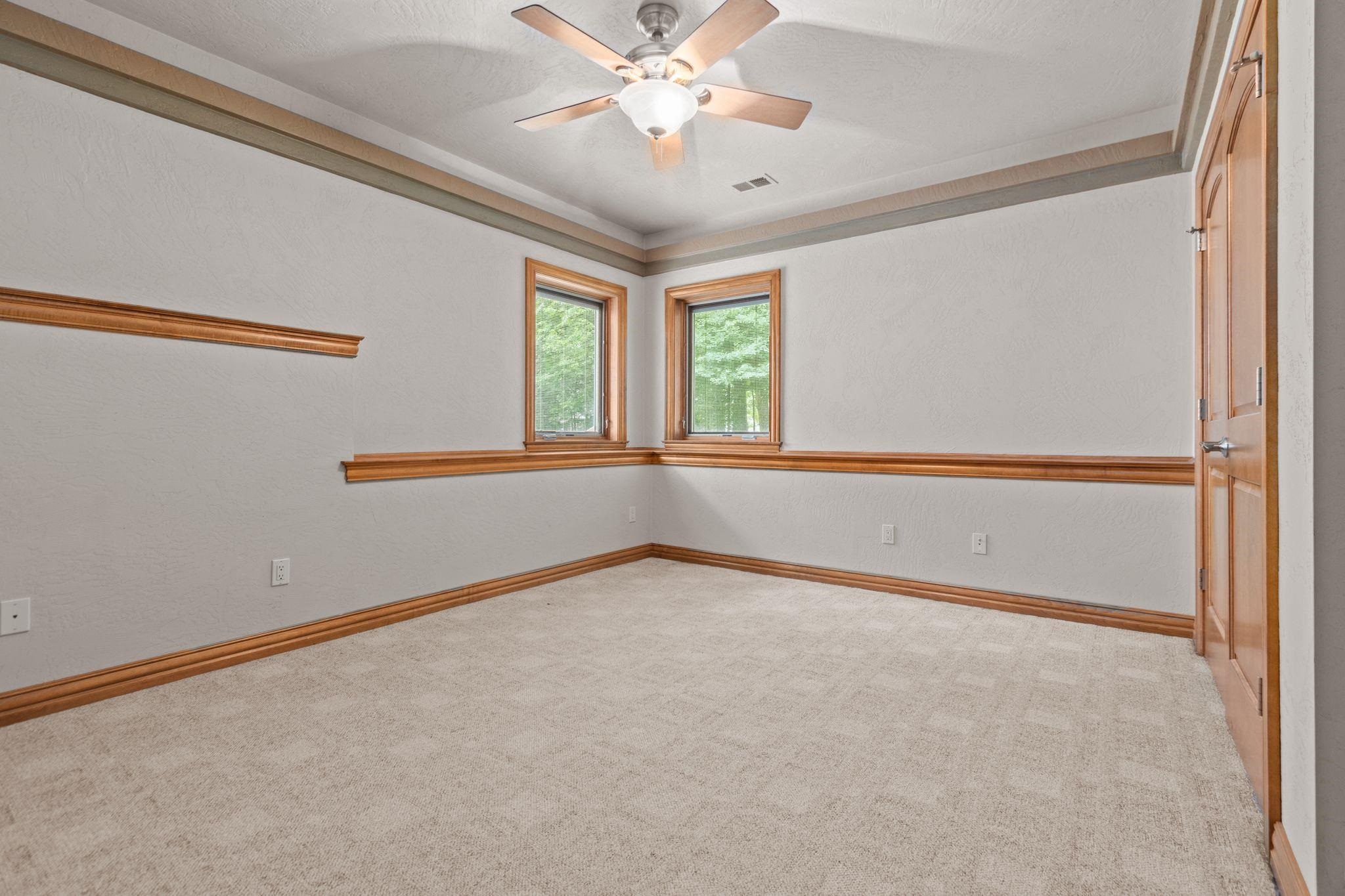 Empty carpeted room with two wood-trimmed windows and a ceiling fan.