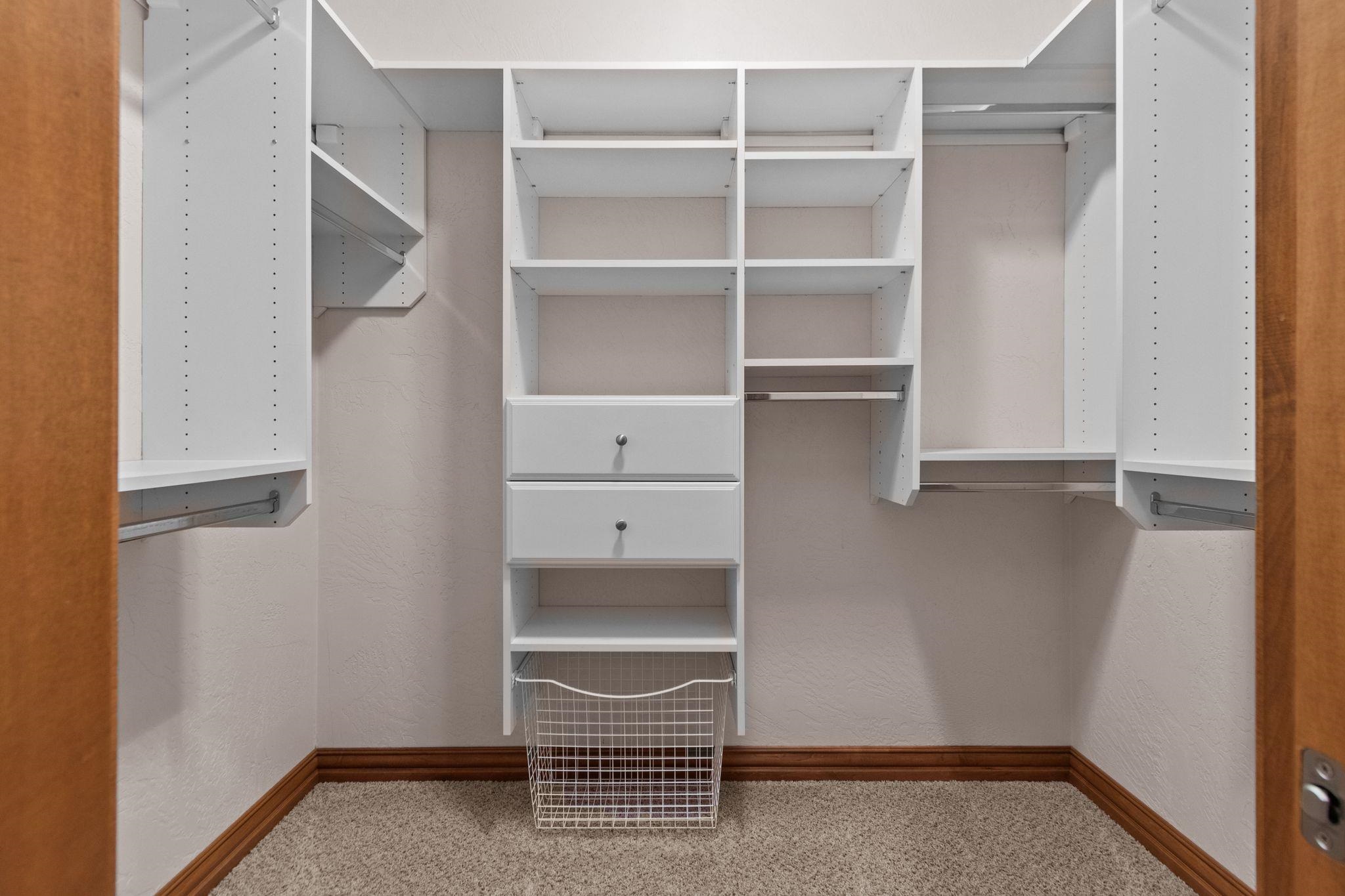 Walk-in closet with white built-in shelving, central drawers, and a wire laundry basket.