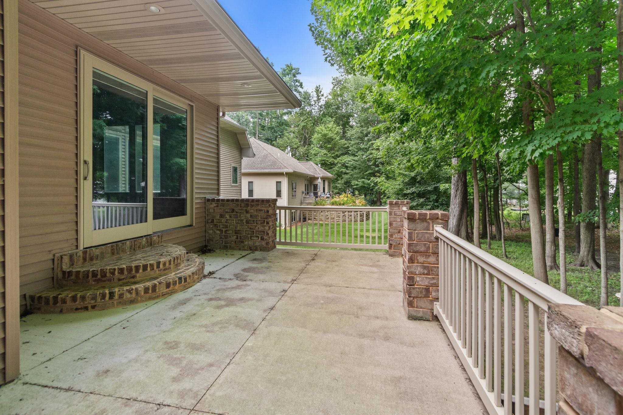 Concrete patio beside a tan-sided home, with curved brick steps and white railings overlooking a leafy yard.