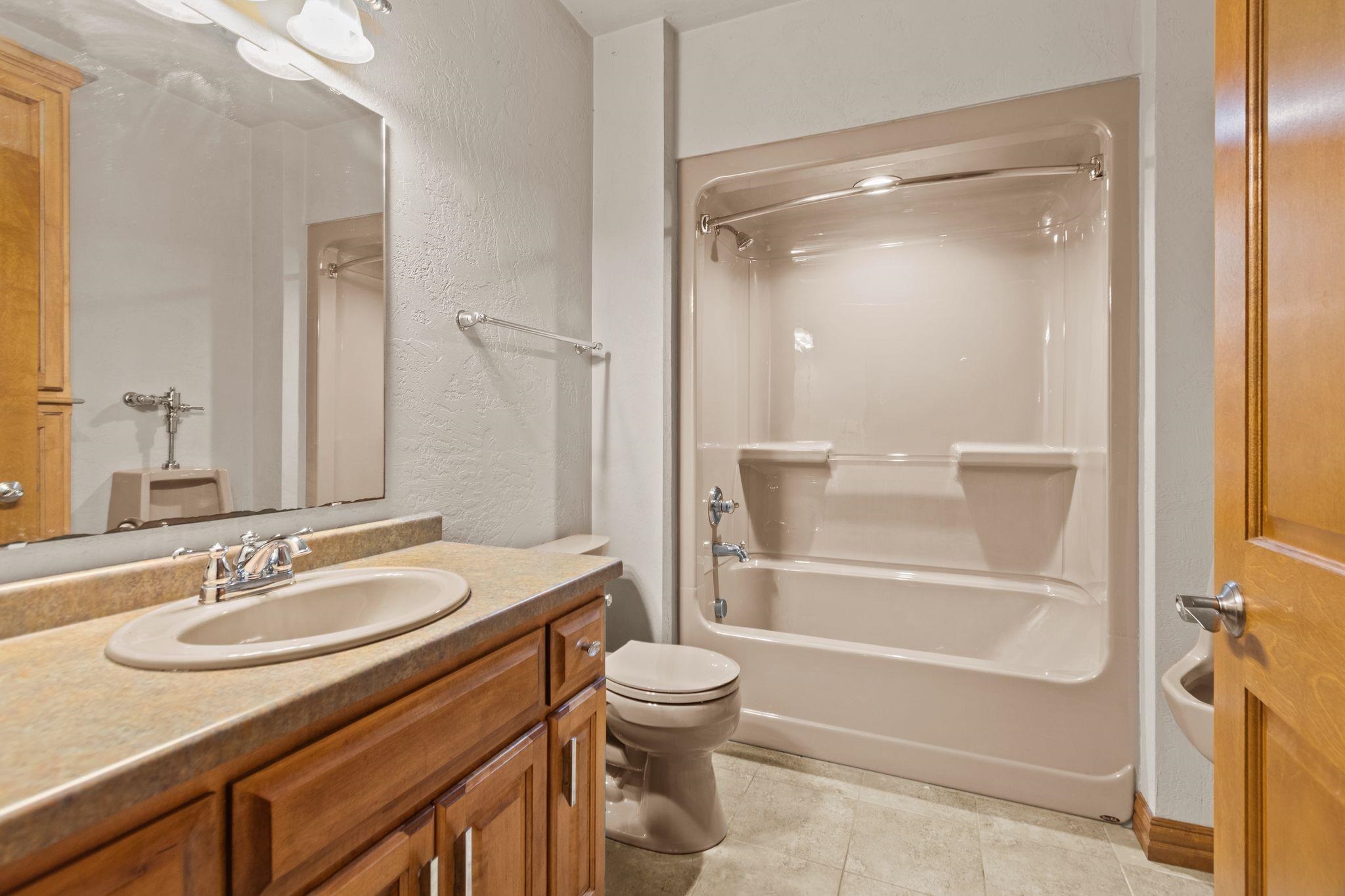 Bathroom with a wood vanity and oval sink beside a toilet and tub-shower with a glass door.