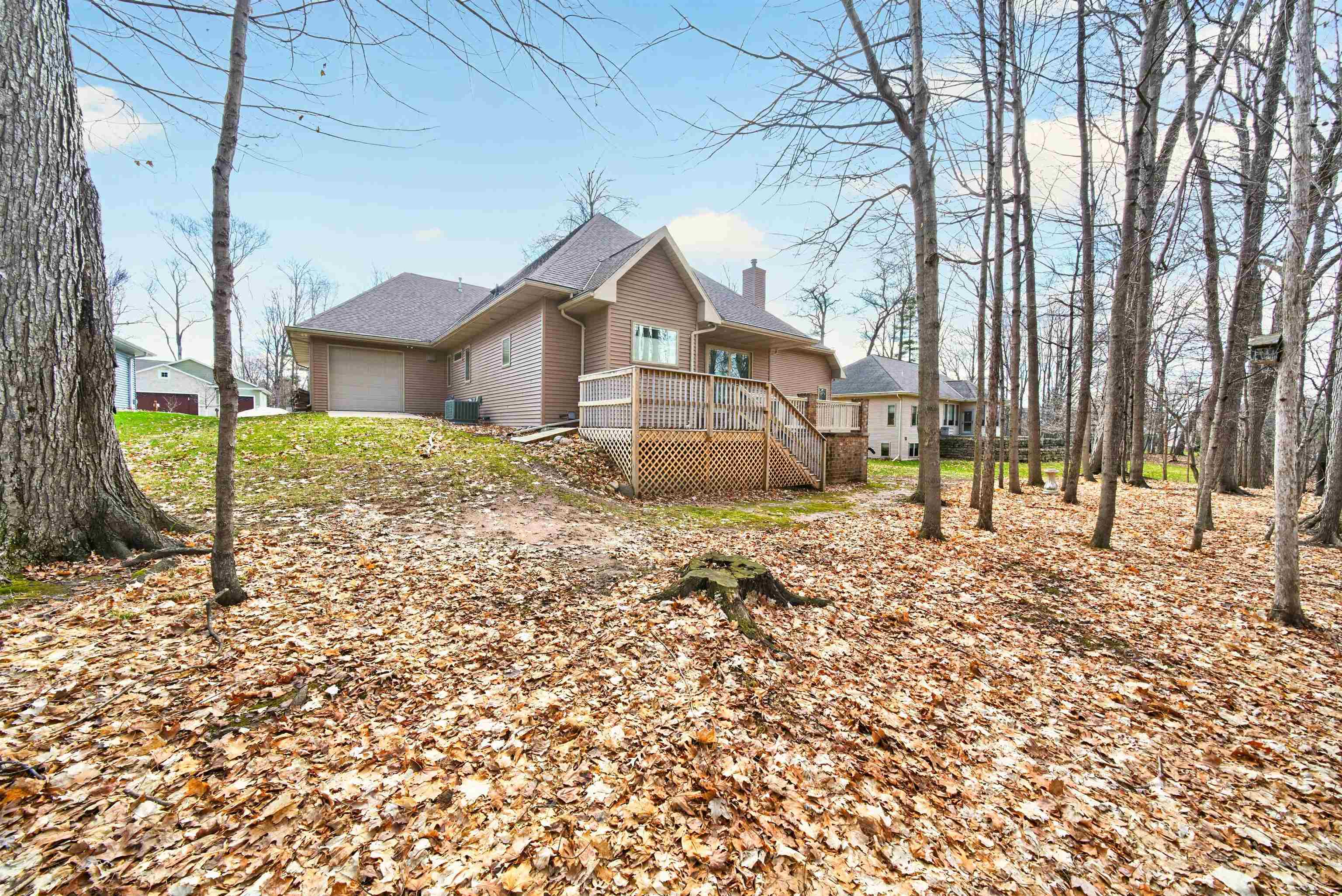 Brown-sided house with an attached garage and raised deck in a wooded, leaf-covered yard.