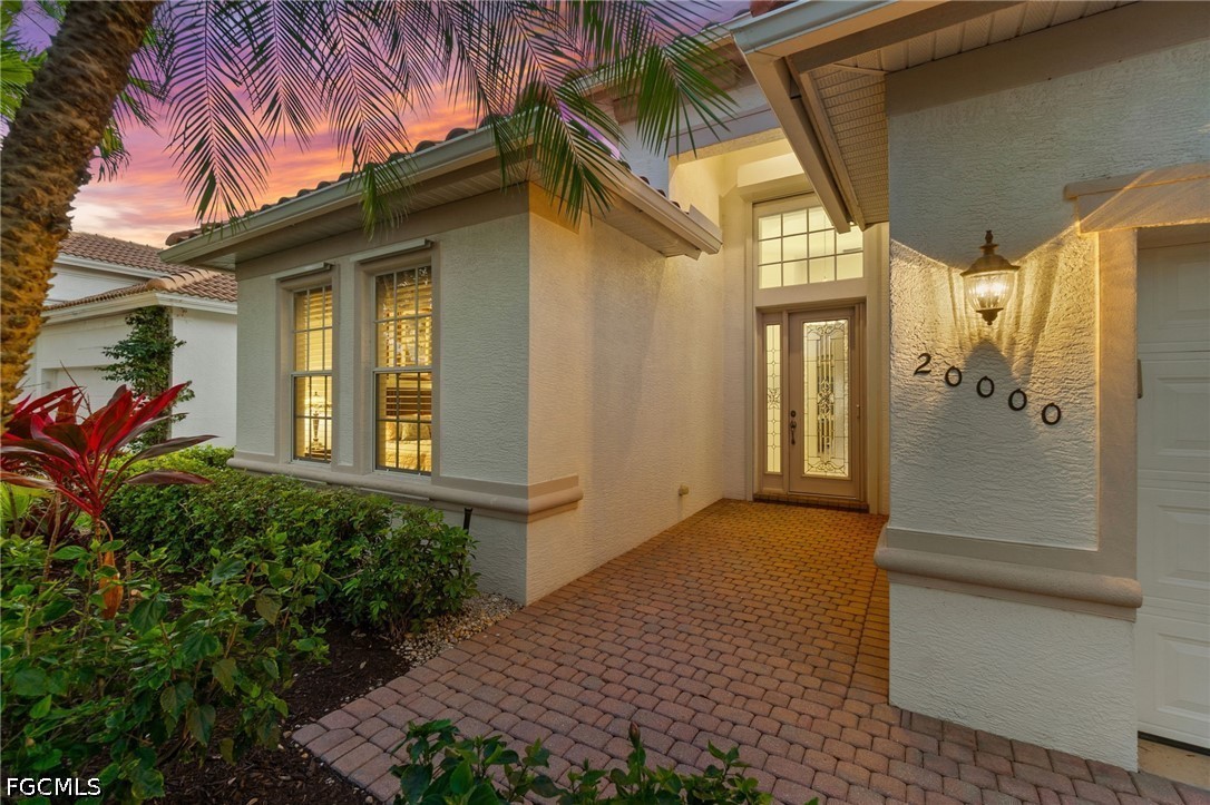 Home entrance with a brick paver walkway, landscaped beds, and glowing glass-paneled door at dusk.