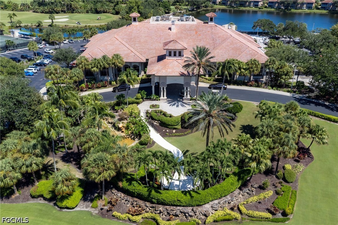 Aerial view of a tiled-roof clubhouse amid palm trees, landscaped grounds, a lake, and golf course.