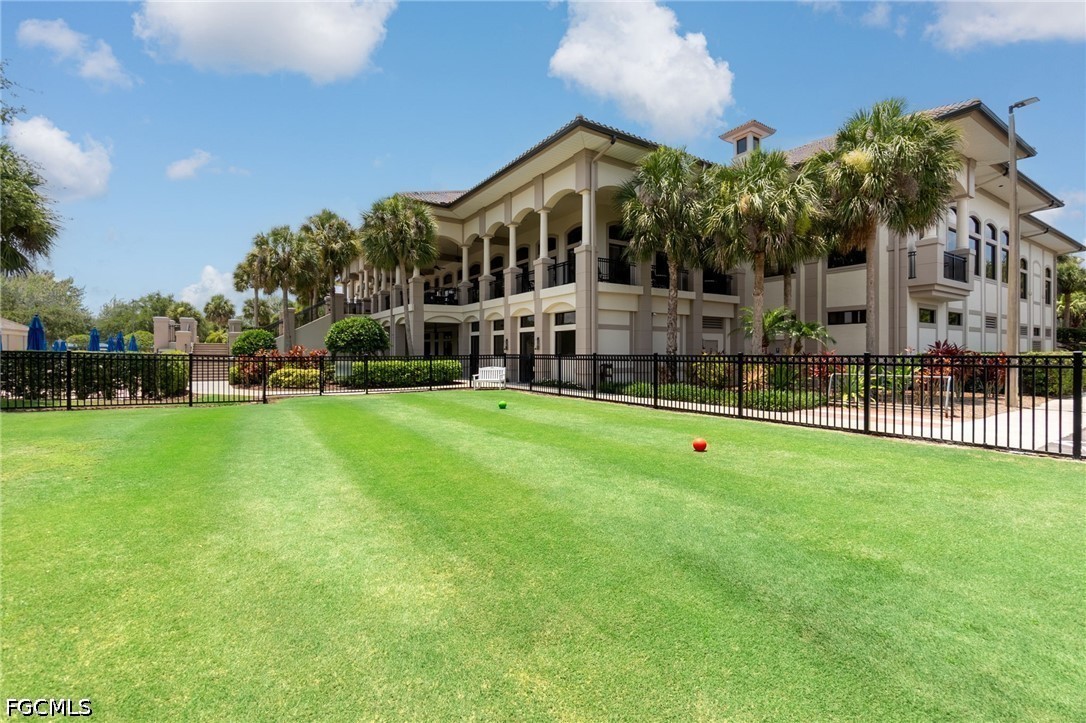 Multi-story beige building with balconies behind a fenced lawn and palm trees.