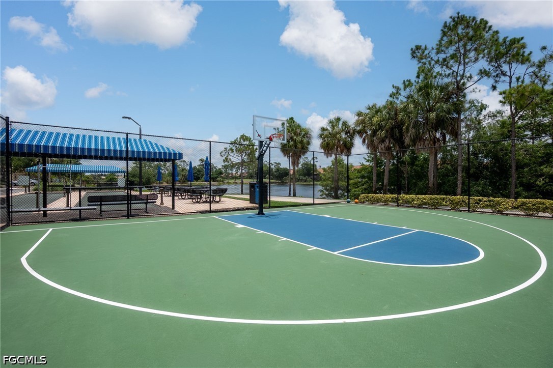Outdoor basketball court with a blue-and-green surface, shaded picnic area, and lake beyond the fence.