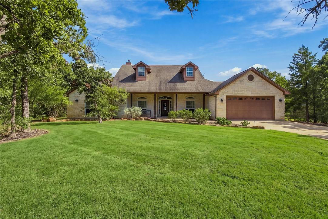 Stone-front house with two dormers and an attached garage, surrounded by a broad lawn and mature trees.