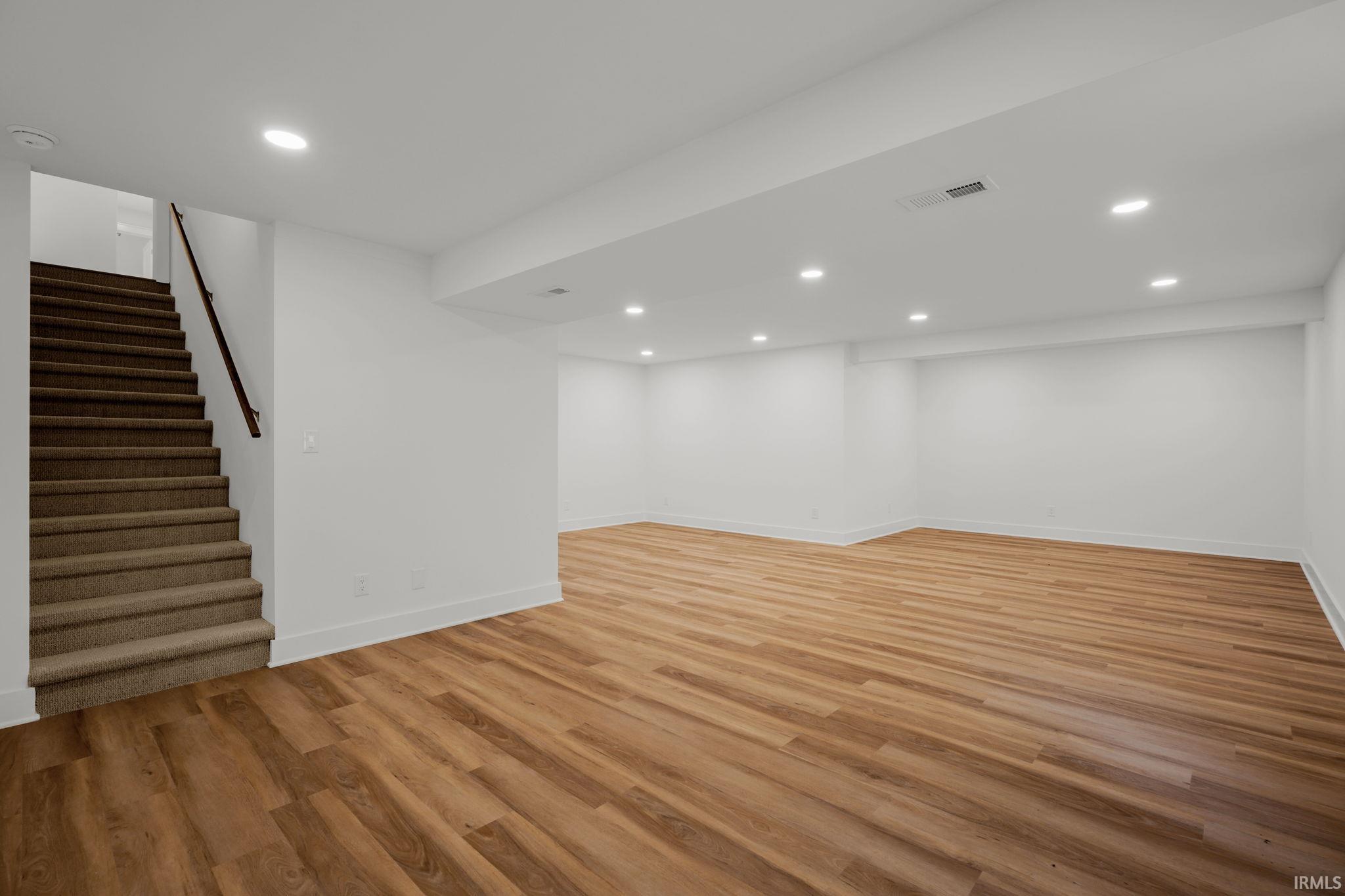 Empty lower-level room with warm wood-look flooring and a carpeted staircase.