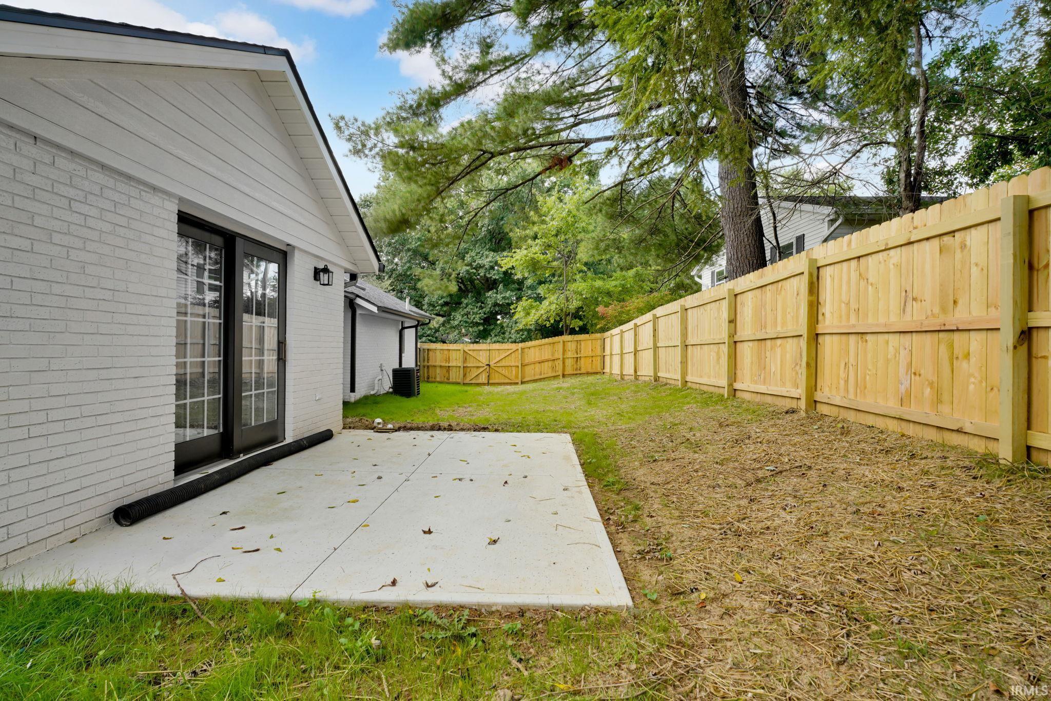 Concrete patio beside a white brick home, with a fenced backyard and mature trees.