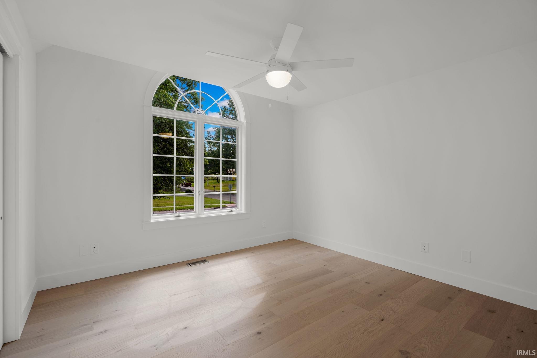 Empty room with pale wood floors and a large arched window overlooking trees and lawn.