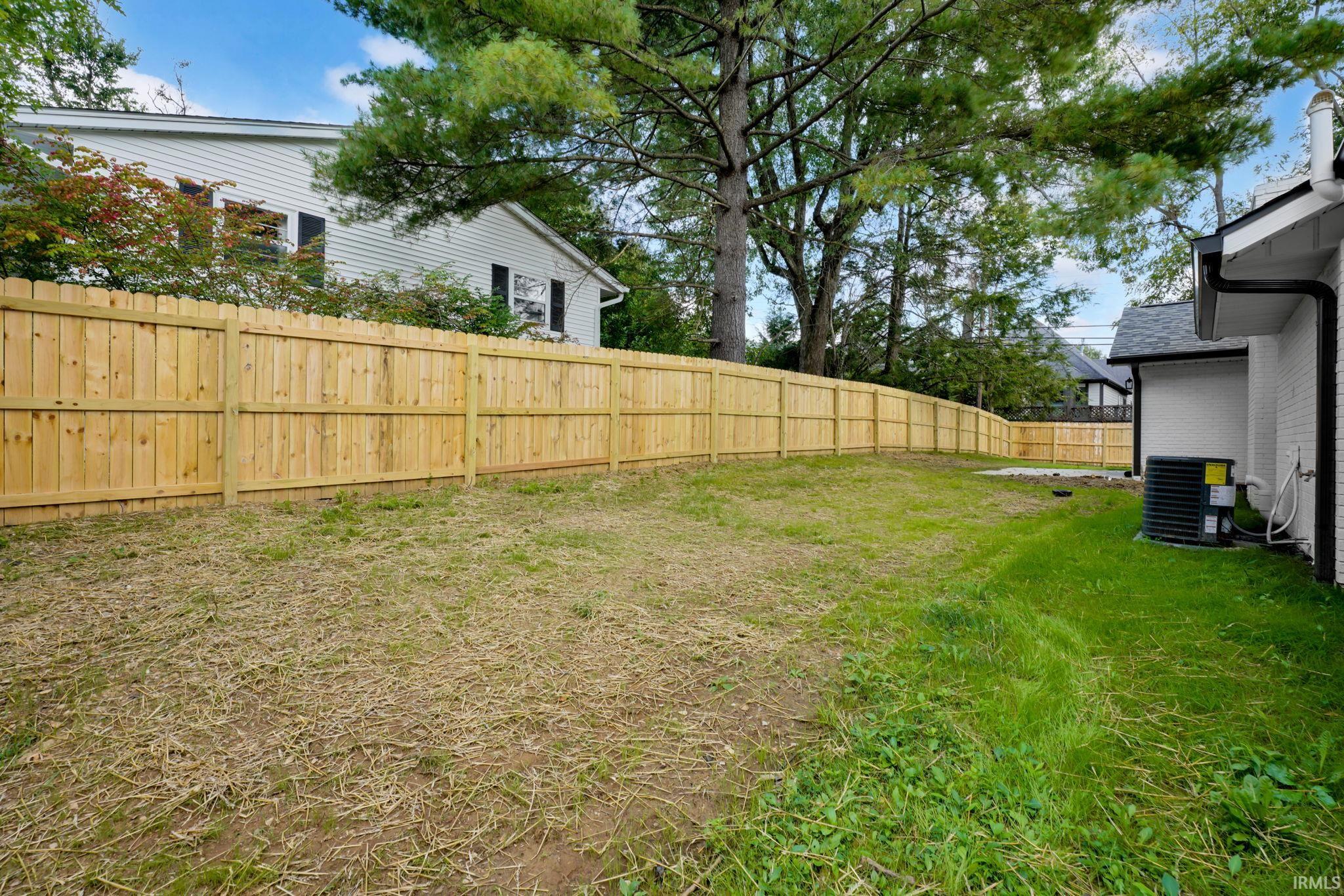Side yard with a long wooden privacy fence, mature trees, and patchy grass.