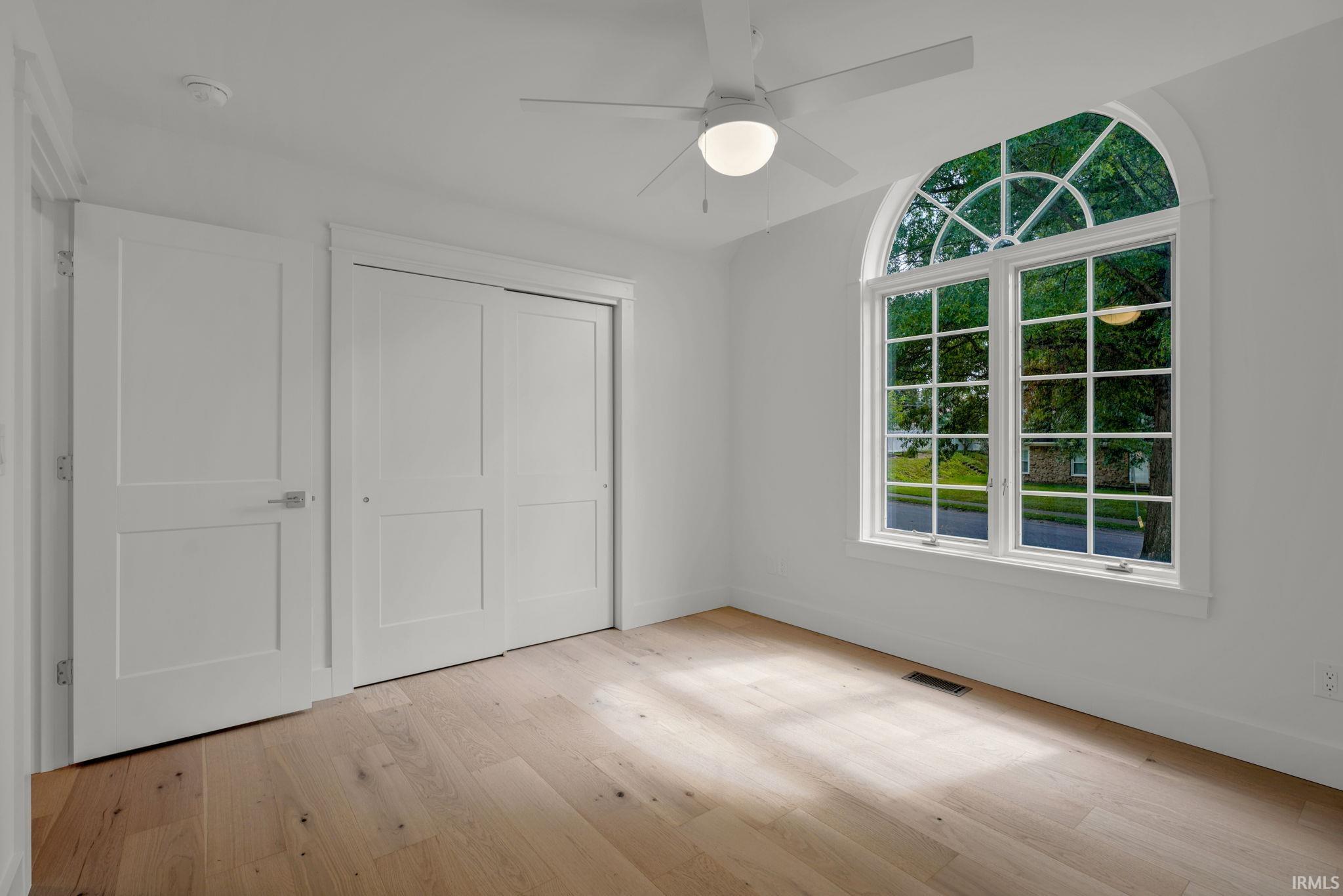 Empty bedroom with pale wood floors, paneled closet doors, and a large arched window.