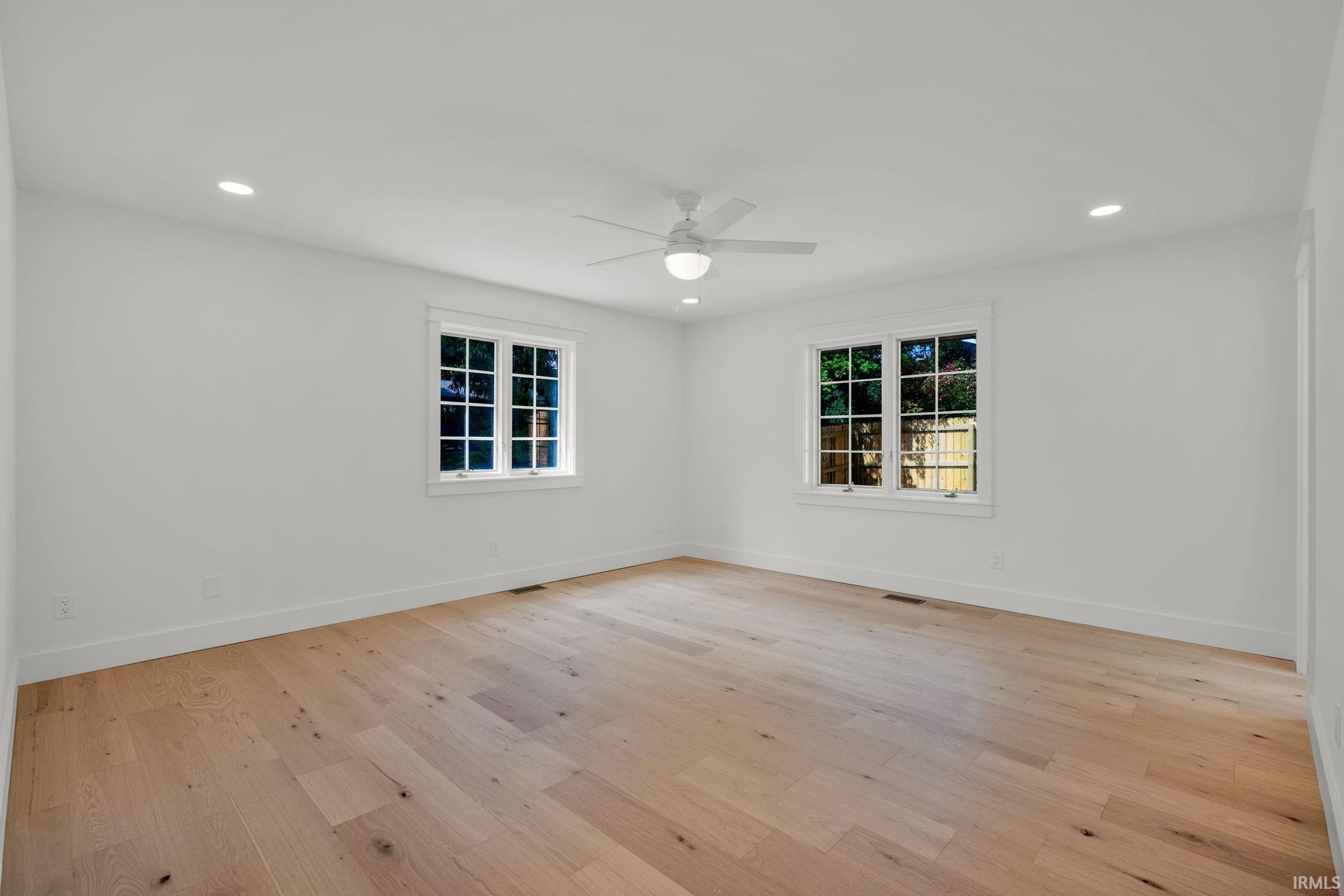 Empty bedroom with pale hardwood floors, two pairs of windows, and a ceiling fan.