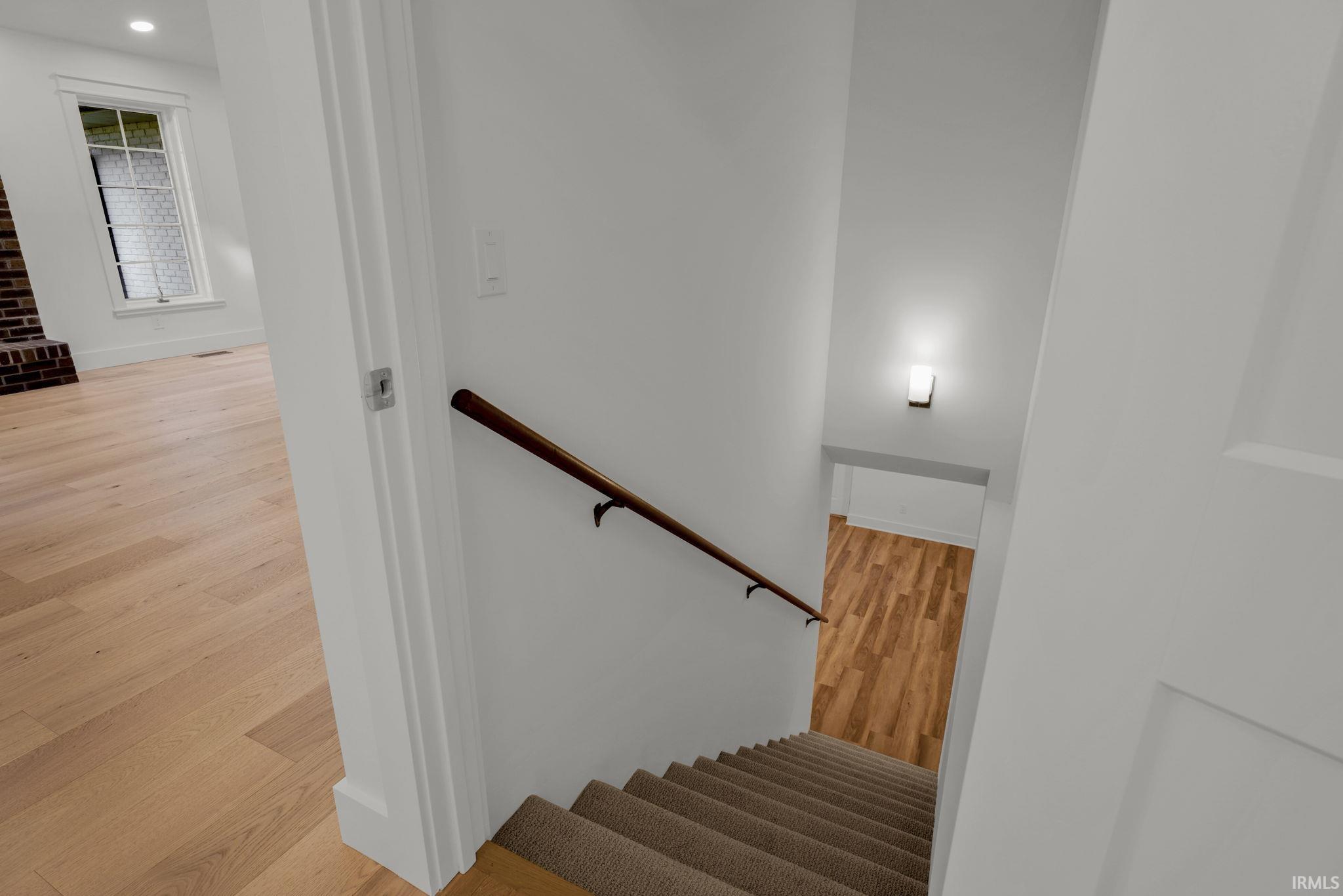 Carpeted staircase with a dark wood handrail beside a bright hardwood-floored room.