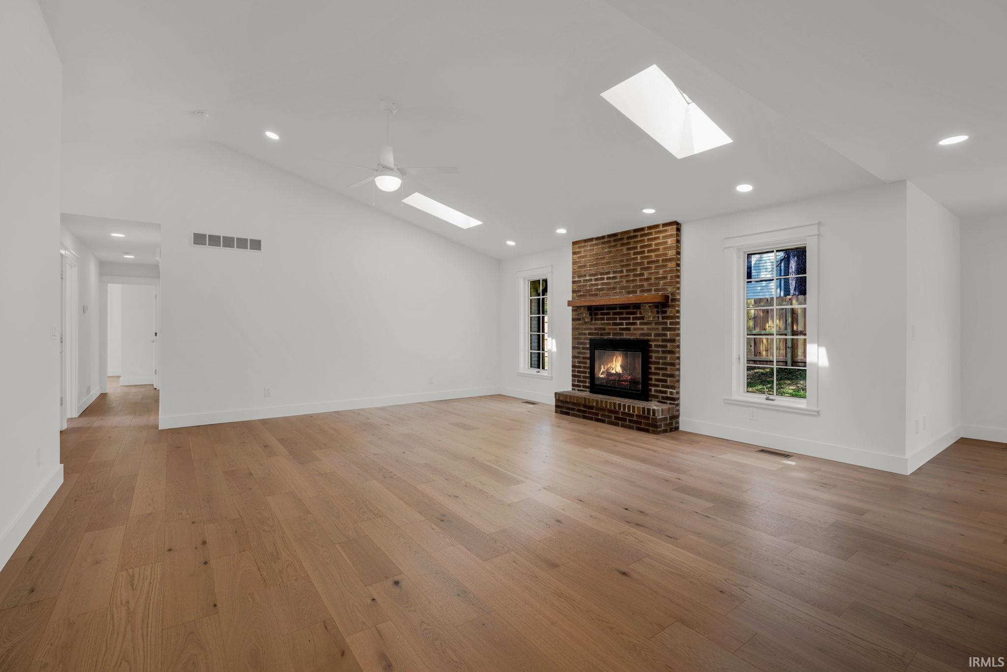 Empty living room with a brick fireplace, light wood floors, and skylights.