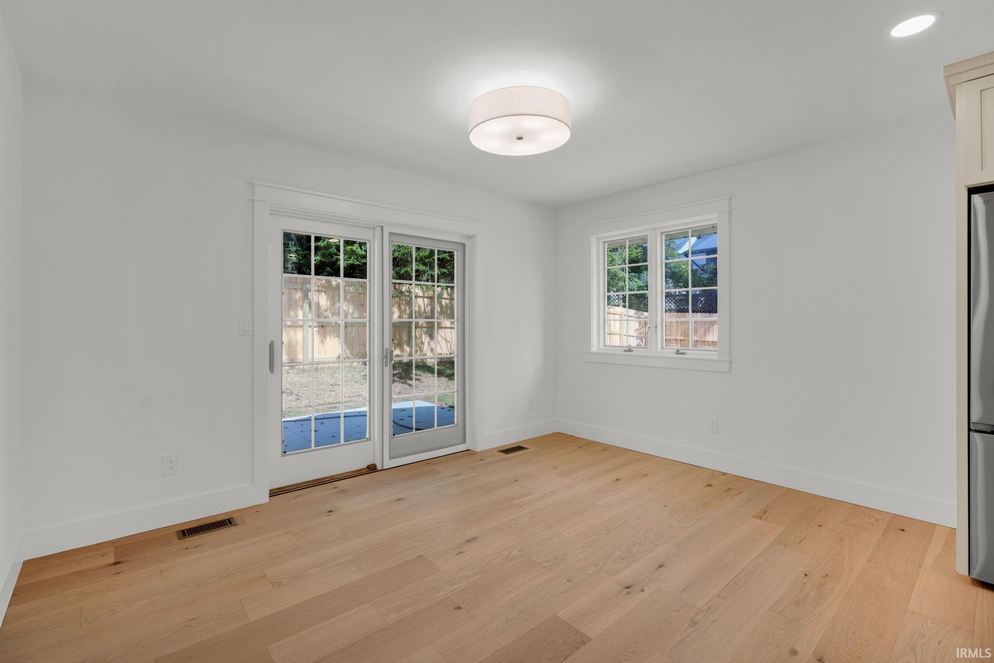 Empty room with light hardwood floors, French doors to a fenced yard, and a large window.