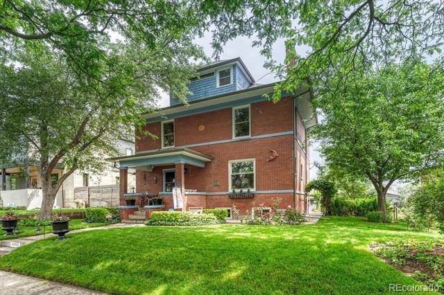 Two-story red-brick house with blue trim and a covered porch, framed by mature trees and a green lawn.