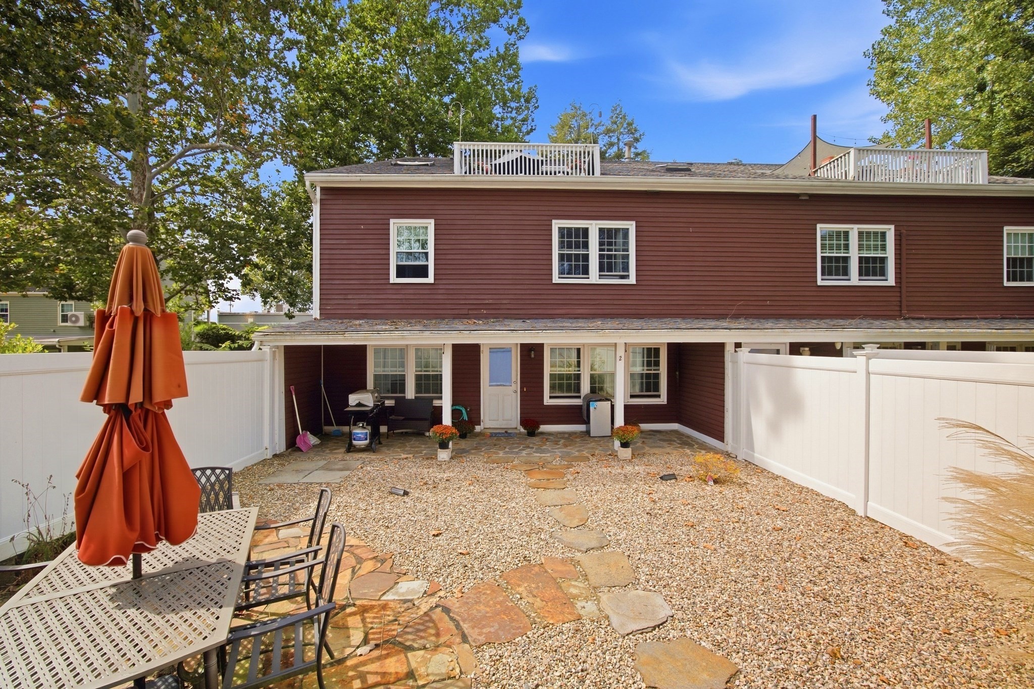 Brown multi-unit home behind a fenced gravel courtyard with a stone path and orange patio umbrella.