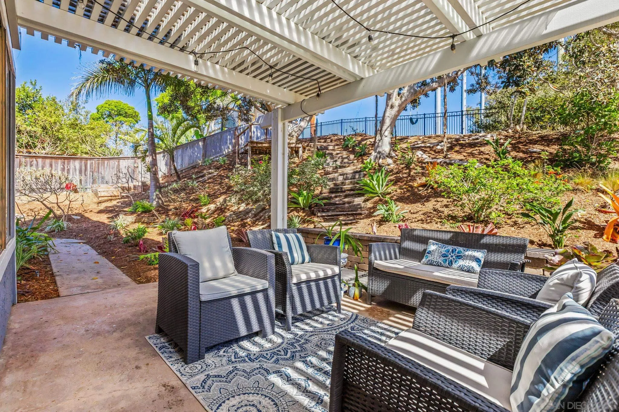 Pergola-covered patio with wicker seating, patterned rug, and steps leading up a landscaped hillside.