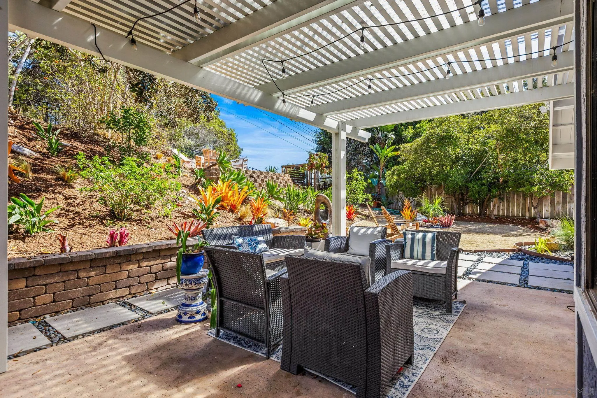 Furnished patio under a white pergola, with wicker seating beside a colorful planted hillside.