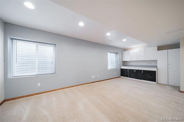 Open-plan room with light carpet, two windows, and a compact kitchen with dark lower cabinets.