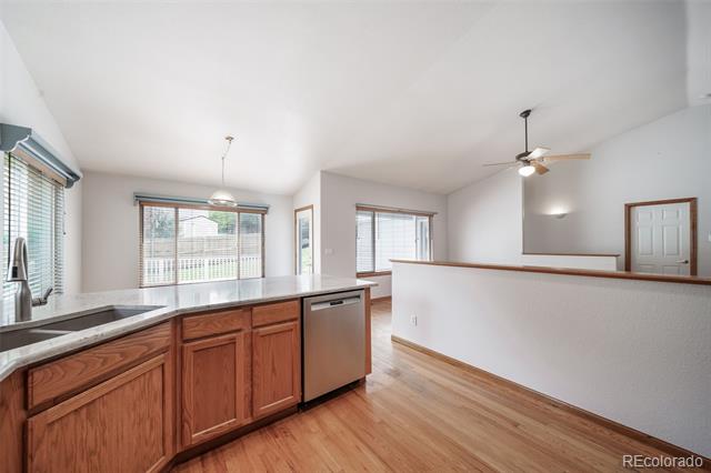 Bright kitchen with wood cabinets, stainless-steel dishwasher, and hardwood floors opening to an unfurnished living area.