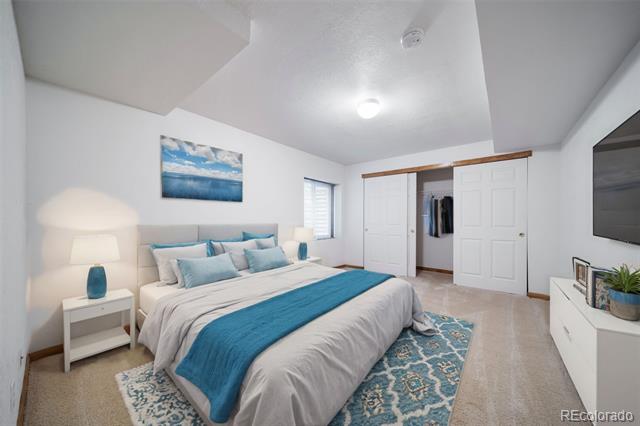 Staged bedroom with a teal bed runner and patterned rug, facing an open closet and wall-mounted television.