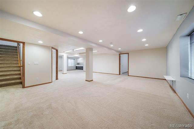 Vacant carpeted room with recessed lighting, a staircase, and a kitchen visible through the doorway.