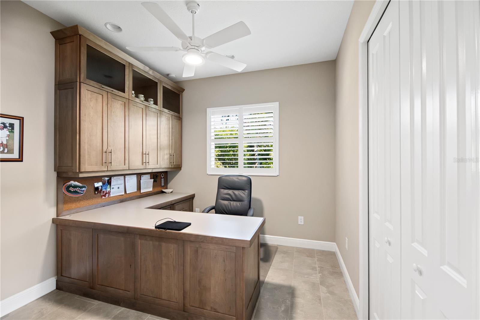 Home office with built-in wood desk and cabinets, black chair, and shuttered window.