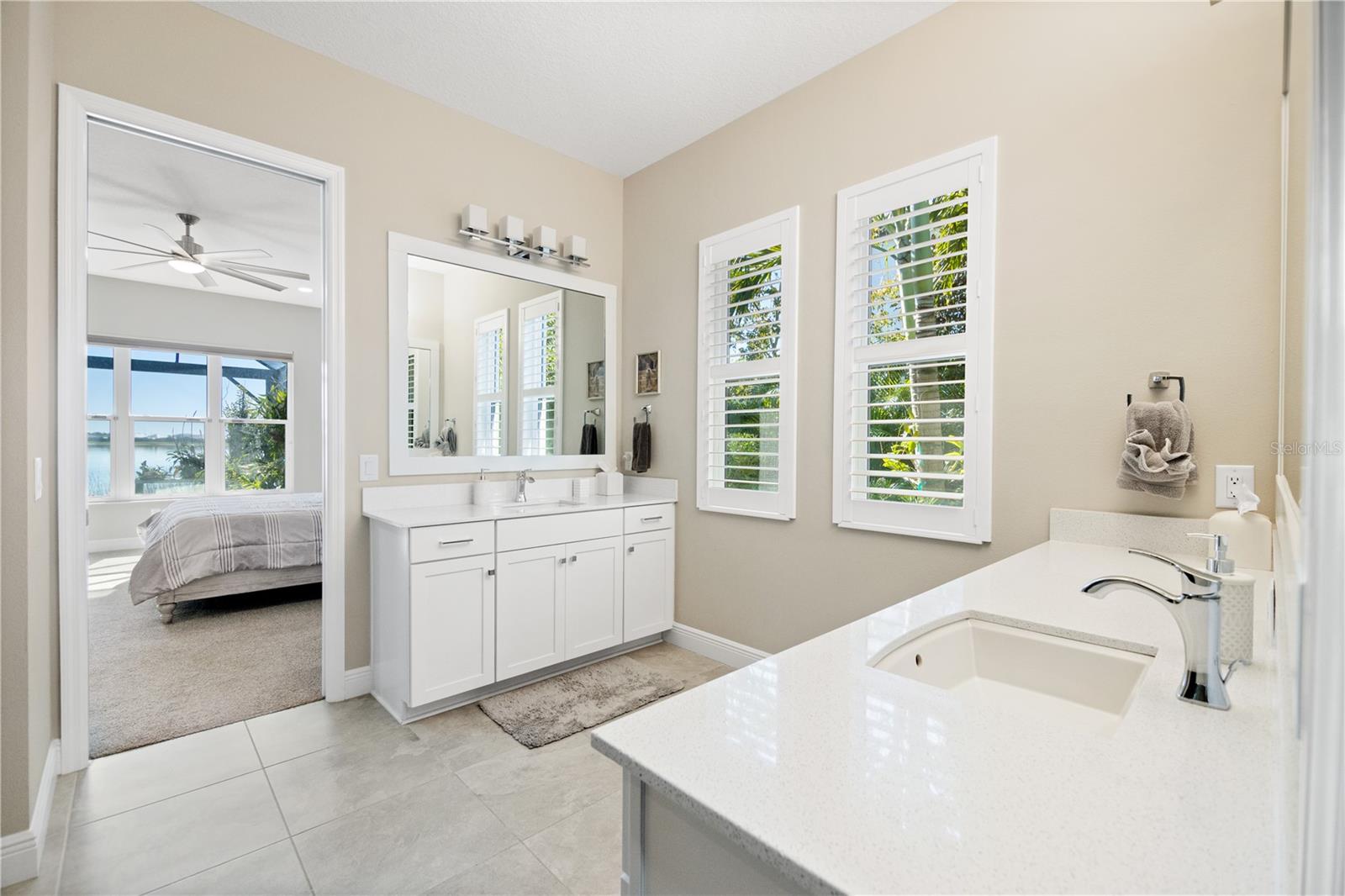 Bright bathroom with white vanities and shuttered windows, opening onto a bedroom with a water view.
