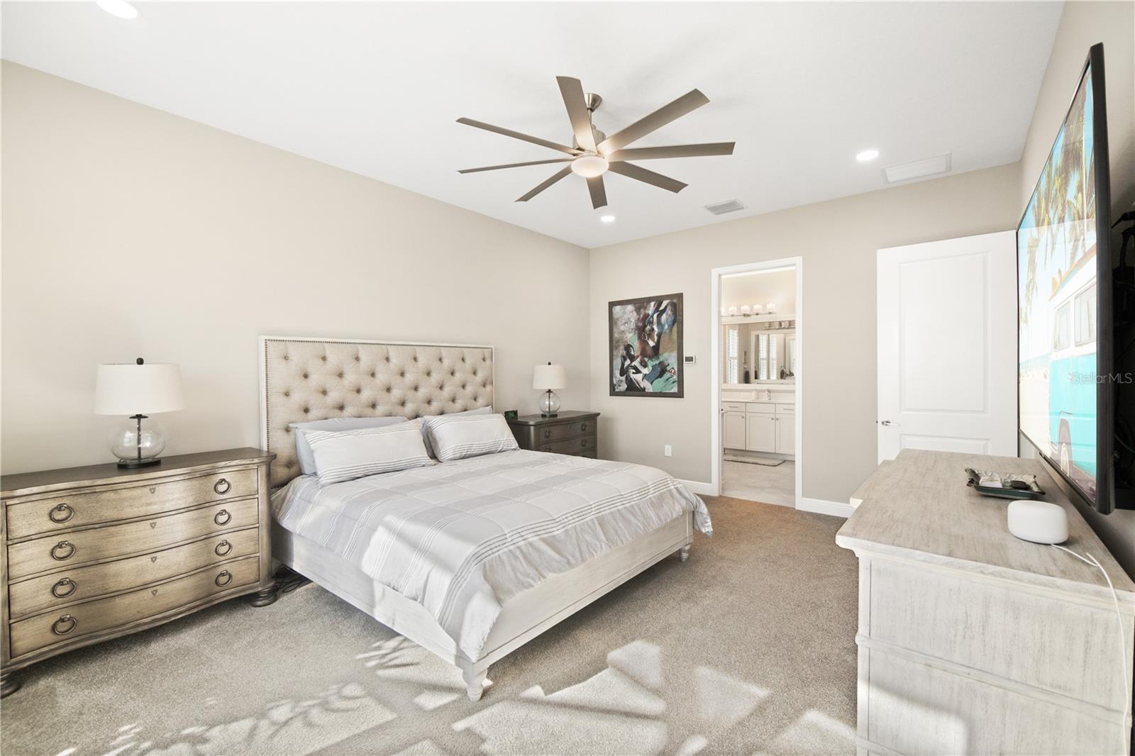 Bedroom with a tufted bed, matching dressers, ceiling fan, and ensuite bathroom doorway.