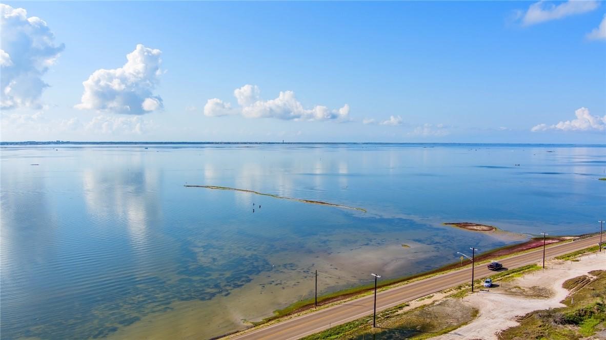 Aerial view of calm coastal water beside a shoreline road under a partly cloudy sky.