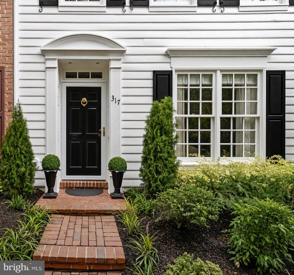 White clapboard home entrance with a black door, curved pediment, and brick walkway through landscaped shrubs.