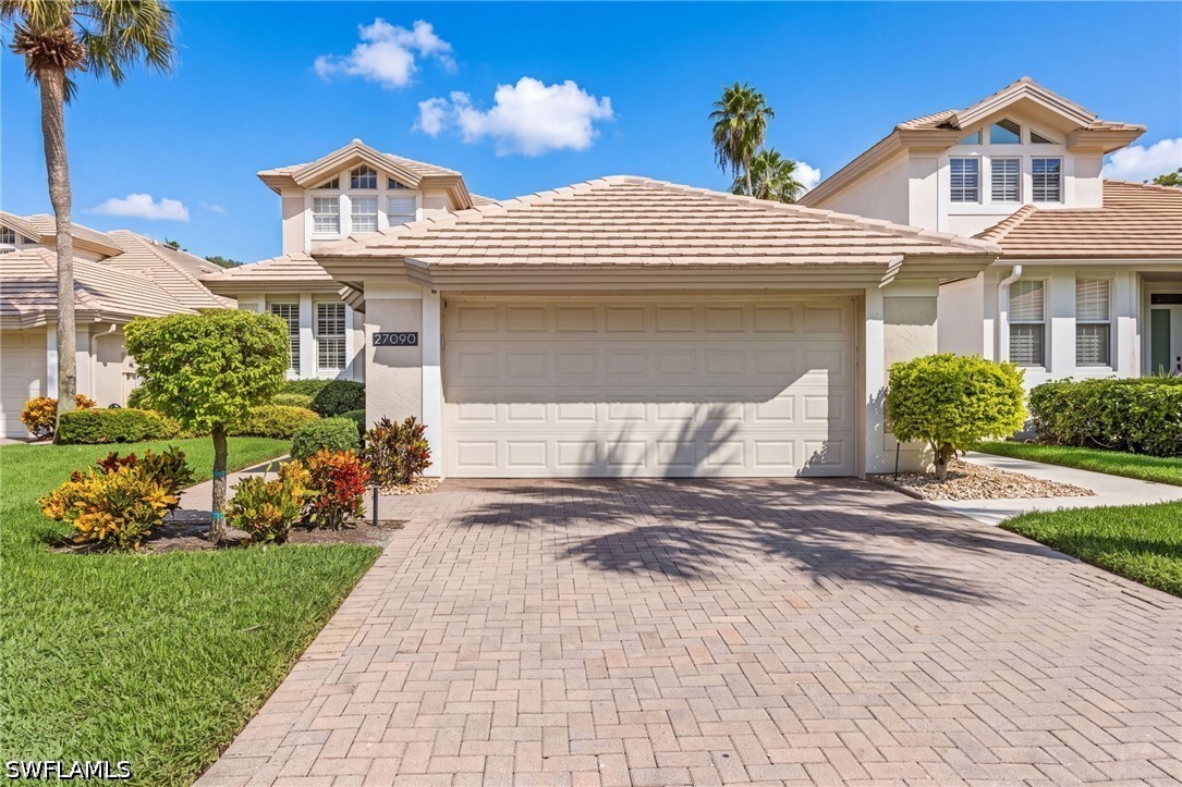 Two-story home with a double garage, broad paver driveway, landscaped lawn, and palm trees.