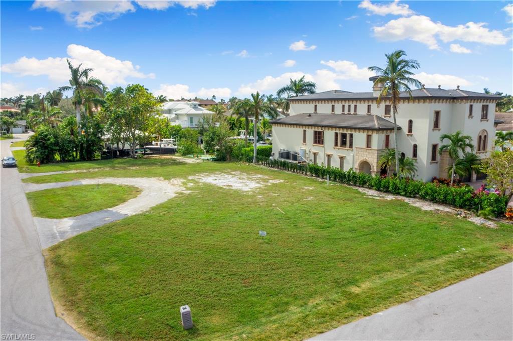 Vacant grassy lot bordered by a road, with a large neighboring home and palm trees.