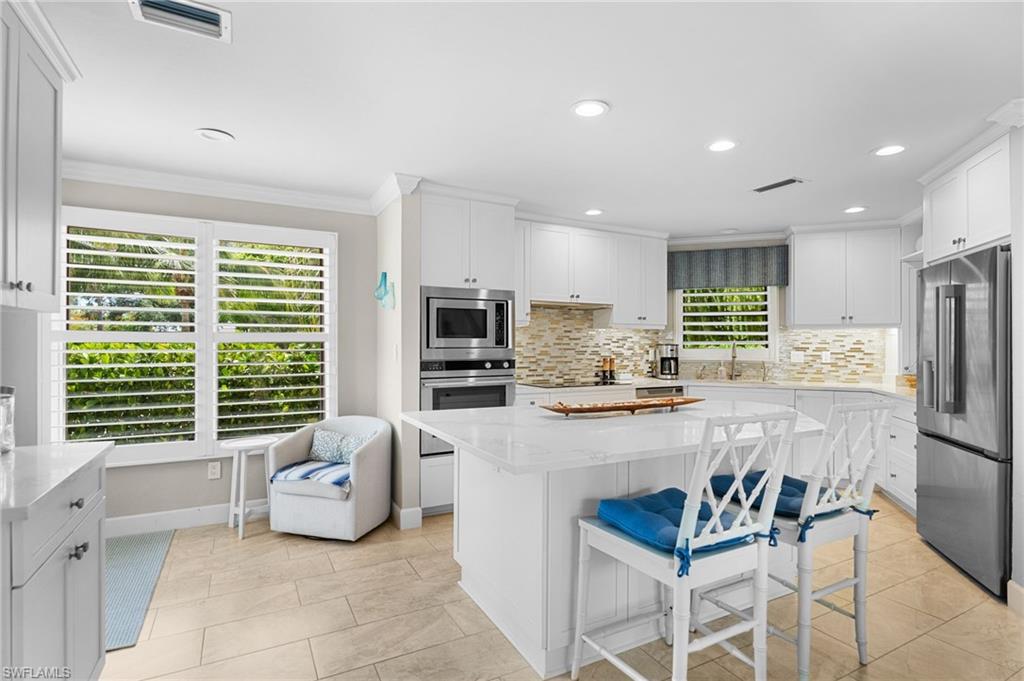 Bright white kitchen with a large island, blue-cushioned stools, and plantation-shuttered windows.
