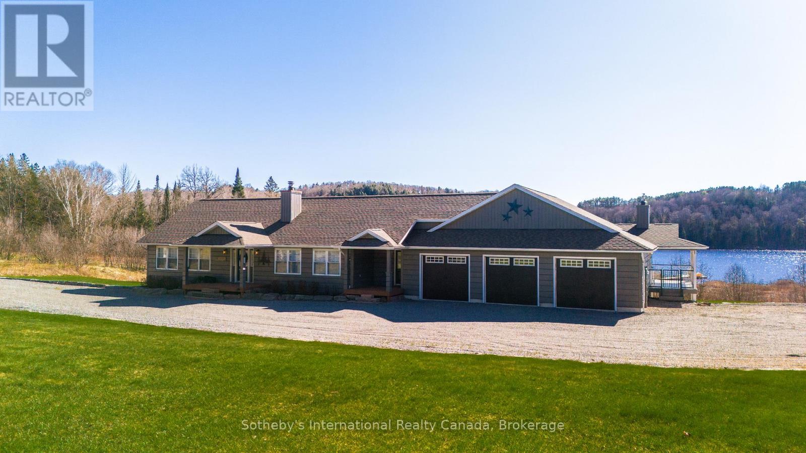 Waterfront home with three-car garage, gravel drive, and wooded hills beyond the lake.