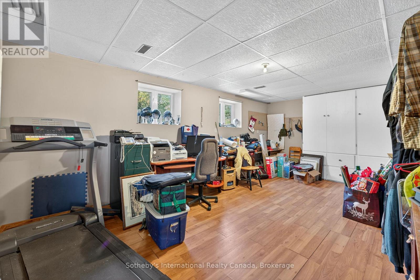 Cluttered room with a treadmill, desk, white storage cabinets, and wood-look flooring.