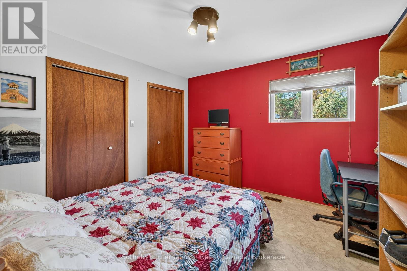 Bedroom with a star-patterned quilt, red accent wall, wooden closets, and a desk by the window.