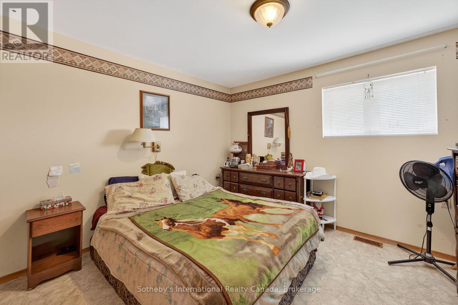 Bedroom with patterned bedding, wooden dresser, and a wide window with blinds.